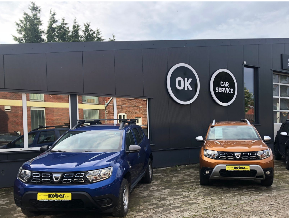 Das Bild zeigt 2 Dacia Duster vor der modernisierten Werkstatt mit dem OK Car Service Logo.